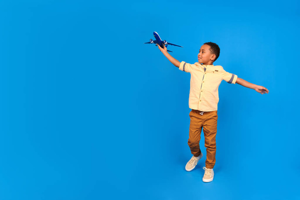 Happy African-American boy plays with a toy plane, puts his hands aside, imagines himself flying in a plane against a blue background. Family travel and tourism, summer holidays concept. - Photo, image