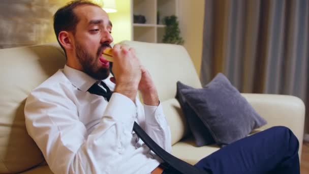 Businessman in formal wear sitting on couch eating a burger - Video