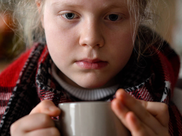 portret van een ziek kind in een sjaal en plaid met een kopje warme thee op de bank in het appartement - Foto, afbeelding