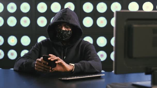 Africano-americano maturo maschio computer di sicurezza hacker indossando maschera viso nero e felpa è seduto in camera moderna scura al desktop, tiene smartphone e lo guarda. - Filmati, video