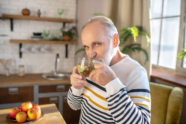 Bearded grey-haired man in a striped sweater having tea in the kitchen - Foto, imagen