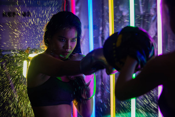 Beautiful 20s Asian Woman trains punching with coach on Silver gold Mitts Gloves. Office Girl exercise at Modern multi color Fashion Neon Muay Thai Boxing Gym with sweat water splash reflect light - Zdjęcie, obraz