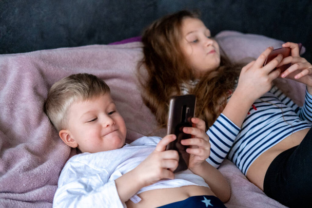 Two kids caucasian boy and girl playing games or looking information video text at Internet on mobile phones at home lying in bed. Digital technology, Modern childhood  - Zdjęcie, obraz