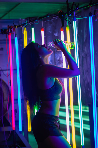 Beautiful 20s Asian Woman finish training in sport bra. Office Girl drinks water from clear bottle through mouth in Modern multi color Fashion Neon Muay Thai Boxing Gym with sweat water splash - Fotoğraf, Görsel