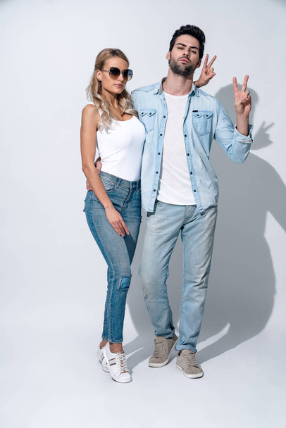Verspieltes Paar. Schönes junges Paar in Jeans macht Friedenszeichen, während es vor grauem Hintergrund steht. - Foto, Bild