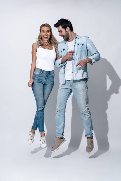 Side view of cheerful modern active couple wearing casual white t shirts and jeans having fun and jumping and dancing, while holding hands and looking at camera on grey background. - Photo, Image