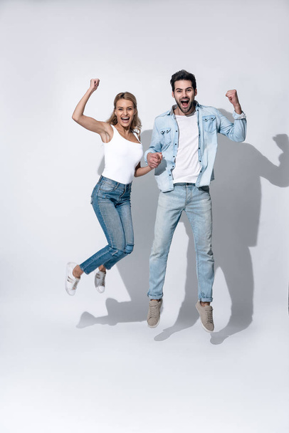 Side view of cheerful modern active couple wearing casual white t shirts and jeans having fun and jumping and dancing, while holding hands and looking at camera on grey background. - Valokuva, kuva