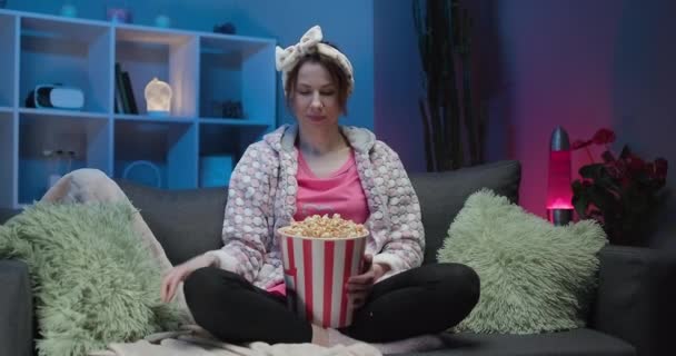 Cheerful woman sitting on the sofa in the living room with popcorn and changing channels with a remote control while watching TV - Video