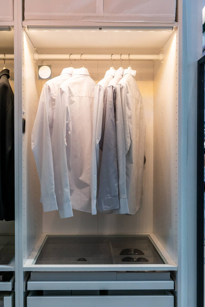Белые и серые основные рубашки висят в шкафу с ящиком для дизайна интерьера
. - Фото, изображение
