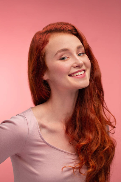 Portrait of smiling redhead girl. Perfect skin. Beautiful female model with long hair over pink background. Young cute woman with clear skin. Skincare and facial treatment concept - Fotografie, Obrázek