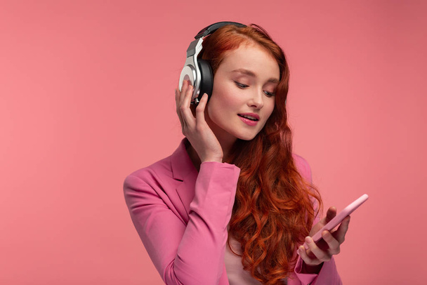 Miłego słuchania muzyki. Piękna, młoda ruda kobieta ze słuchawkami słuchającymi muzyki na smartfonie za pomocą aplikacji muzycznej. Zabawna uśmiechnięta dziewczyna ze słuchawkami i telefonem komórkowym na różowym tle - Zdjęcie, obraz