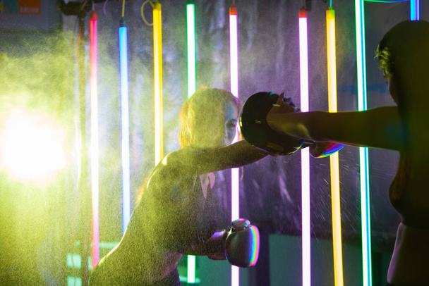 Όμορφη εικοσάρα ασιατική γυναίκα τρένα punching με πούλμαν σε ασημένια χρυσά γάντια γάντια. Office Girl άσκηση στο σύγχρονο πολύχρωμο Μόδα Neon Muay Thai Πυγμαχία Γυμναστήριο με νερό ιδρώτα splash αντανακλούν το φως - Φωτογραφία, εικόνα