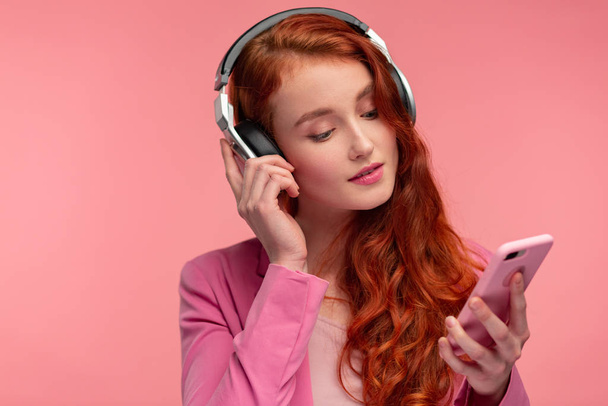 Enjoy listening to music. Beautiful young redhead woman with headphones listening music on smart phone using music app. Funny smiling girl with earphones and mobile phone on pink background - Foto, imagen