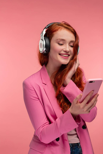 Enjoy listening to music. Beautiful young redhead woman with headphones listening music on smart phone using music app. Funny smiling girl with earphones and mobile phone on pink background - Photo, image