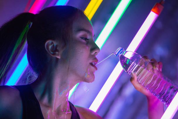 Beautiful 20s Asian Woman finish training in sport bra. Office Girl drinks water from clear bottle through mouth in Modern multi color Fashion Neon Muay Thai Boxing Gym with sweat water splash - Zdjęcie, obraz