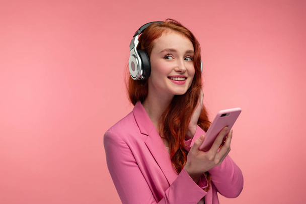 Enjoy listening to music. Beautiful young redhead woman with headphones listening music on smart phone using music app. Funny smiling girl with earphones and mobile phone on pink background - 写真・画像