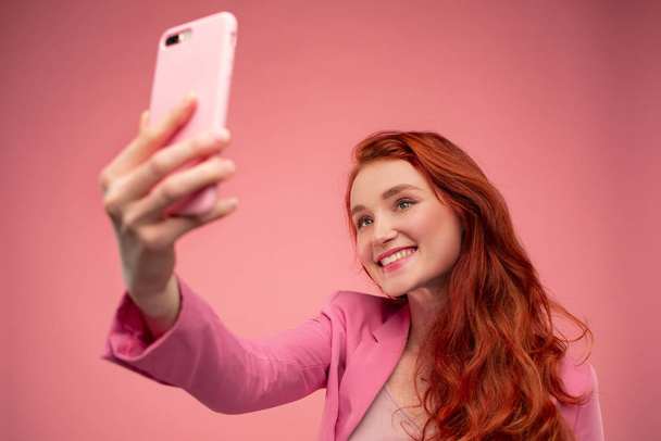 Beautiful young redhead woman making selfie. Funny smiling girl wearing pink jacket holding pink smartphone and posing for selfie isolated on pink background - Zdjęcie, obraz