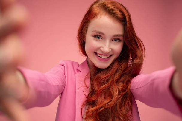 Close-up portrait of beautiful young redhead woman making selfie using two hands. Funny smiling girl wearing pink jacket giving a gift isolated on pink background. Copy space - 写真・画像