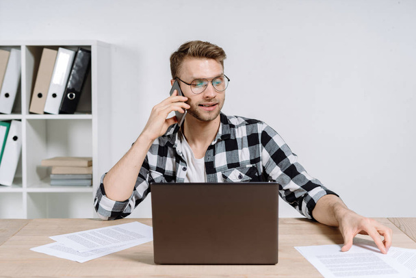 Концентрований і серйозний молодий менеджер дорослих сидить в офісі за сучасним ноутбуком, перевіряє документи, розмовляє з клієнтом на смартфоні
 - Фото, зображення