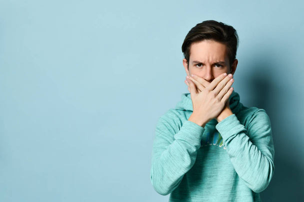 Young teen boy in blue hoodie standing and closing mouth to keep silence over pastel blue wall background - Foto, Bild