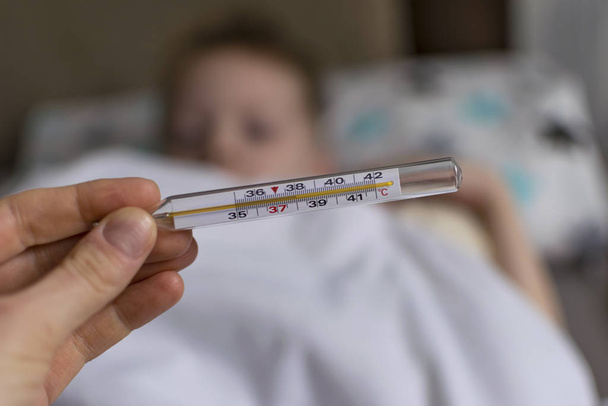 Ateşi ve baş ağrısı olan hasta üzgün bir çocuk yatakta yatıyor, bir termometre. Grip salgını virüsü bakterisi - Fotoğraf, Görsel