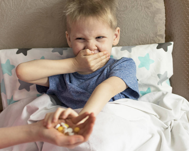 Ateşi ve baş ağrısı olan hasta üzgün bir çocuk yatakta yatıyor, ilaç ve hap içmeyi reddediyor. Grip salgını virüsü bakterisi - Fotoğraf, Görsel