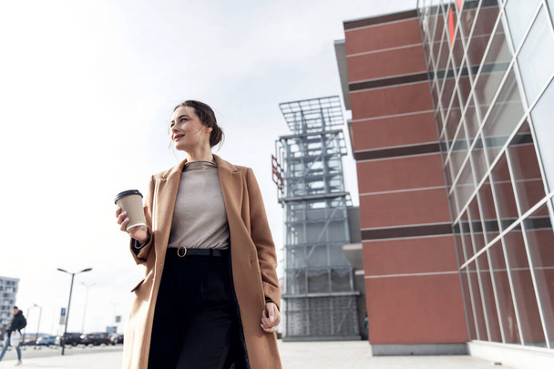 Young cousian woman with smartphone standing against street blurred building background. Fashion business photo of beautiful girl in black casual suite with phone and cup of coffee - Fotoğraf, Görsel