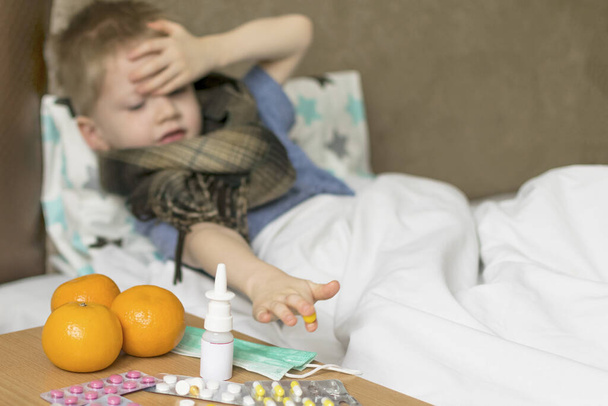 Ένα άρρωστο θλιμμένο παιδί με πυρετό και πονοκέφαλο βρίσκεται στο κρεβάτι δίπλα στα φάρμακα. βακτήριο του ιού της νόσου των κρυολογημάτων γρίπης - Φωτογραφία, εικόνα