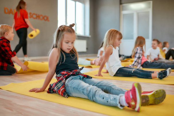 Portrait of a pretty little girl sitting on the yellow yoga mat while having a yoga class in the dance studio. Children doing gymnastic exercises - Foto, afbeelding