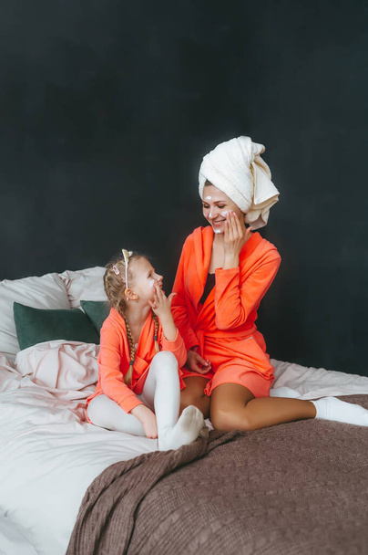 Ευτυχισμένη μητέρα και η κόρη της με μπουρνούζια και μάσκα κρέμας στο κρεβάτι στο σπίτι. Οικογενειακή φροντίδα και σπα - Φωτογραφία, εικόνα