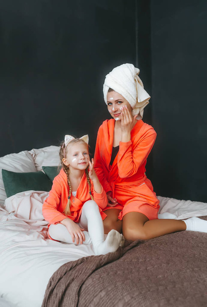 Gelukkige moeder en dochter in badjassen met crème masker in bed. Familieportret.  - Foto, afbeelding