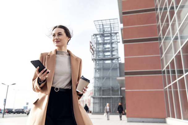 スマートフォンを背景に立っている若い女性がビルをぼやけている。ファッションビジネス写真の美しい女の子で黒カジュアルなスイートで電話とコーヒーカップ - 写真・画像