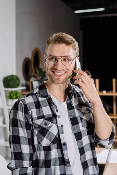 Retrato de un joven freelancer feliz parado en el lugar de trabajo, hablando en un smartphone moderno con el cliente, haciendo sonreír a los dientes, mirando a un lado
 - Foto, imagen