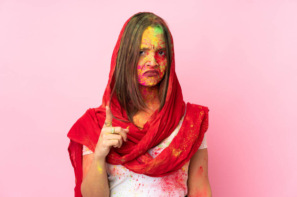 Mujer india joven con coloridos polvos de holi en su cara aislada sobre fondo rosa frustrado y apuntando hacia el frente
 - Foto, Imagen