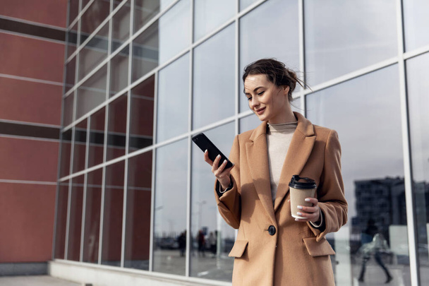 Closeup young Business Woman holding smartphone, Coffee to go and smiling. Attractive stylish Woman using her Phone near modern Building. Having Break. Smartphones. Games. People. Emotions. Business. - Fotó, kép