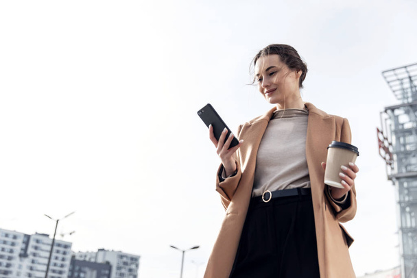 Closeup young Business Woman holding smartphone, Coffee to go and smiling. Attractive stylish Woman using her Phone near modern Building. Having Break. Smartphones. Games. People. Emotions. Business. - Photo, Image