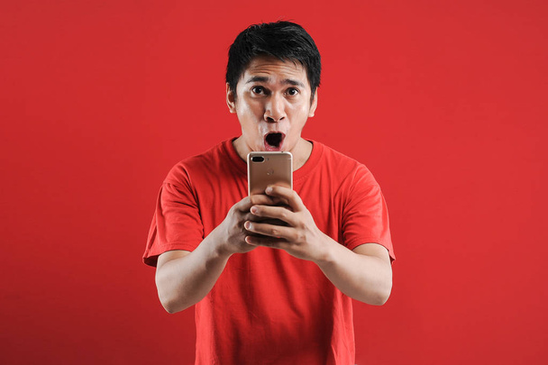 Happy Asian man reading new text on mobile, isolated on red background - Photo, Image