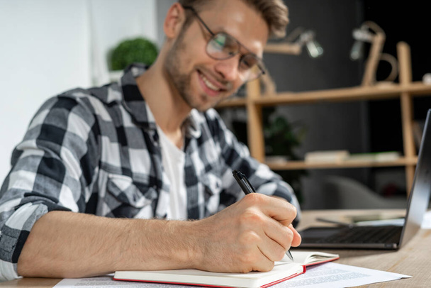 Вибірковий фокус на успішному молодому бізнесмену, який пише плани в організаторі, сидячи на робочому місці за столом і роблячи блимаючу посмішку
 - Фото, зображення