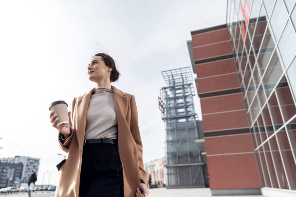 Young cousian woman with smartphone standing against street blurred building background. Fashion business photo of beautiful girl in black casual suite with phone and cup of coffee - Fotografie, Obrázek