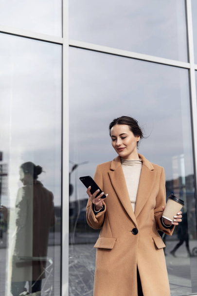 Business Woman smiling charmingly, using her Smartphone. Female enjoying sunny day and funny conversation. Typing on her Phone with Interest. Brunette Girl. Successful Woman. Phones. Apps. - Zdjęcie, obraz