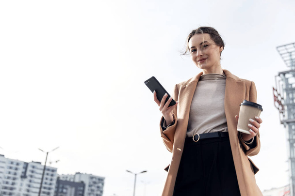 Closeup young Business Woman holding smartphone, Coffee to go and smiling. Attractive stylish Woman using her Phone near modern Building. Having Break. Smartphones. Games. People. Emotions. Business. - Foto, Imagen