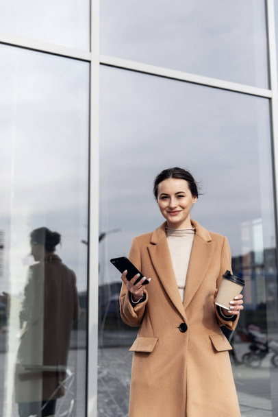 Closeup young Business Woman holding smartphone, Coffee to go and smiling. Attractive stylish Woman using her Phone near modern Building. Having Break. Smartphones. Games. People. Emotions. Business. - Photo, image