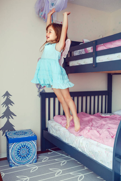 Cute Caucasian girl jumping from bed. Happy excited kid having fun at home. Adorable child playing game flying like elf or fairy. Authentic action candid lifestyle domestic life moment.  - Foto, imagen