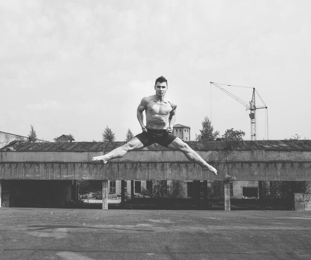 Νεαρός όμορφος άνδρας με γυμνό κορμό χορευτής πήδηξε ψηλά στον αέρα σε βιομηχανικό κτίριο. Σύγχρονη τέχνη χορό. Ασπρόμαυρο. Οδός αστικός τρόπος ζωής. - Φωτογραφία, εικόνα