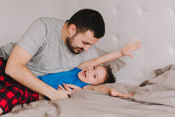 Young Caucasian father having fun with boy son. Man parent hugging with child on bed in bedroom at home. Authentic lifestyle real candid family moment. Happy Fathers Day holiday.  - 写真・画像