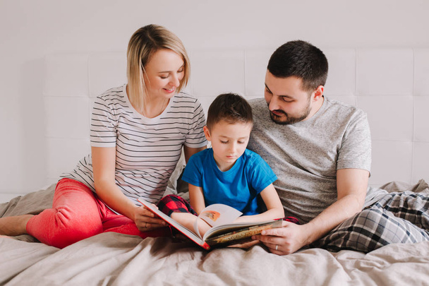 Family of three people sitting on bed in bedroom reading book. Mother, father and boy son at home spending time together. Parents talking communicate to child. Real people authentic lifestyle  - Photo, Image