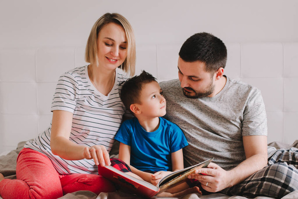 Family of three people sitting on bed in bedroom reading book. Mother, father and boy son at home spending time together. Parents talking communicate to child. Real people authentic lifestyle  - Фото, изображение