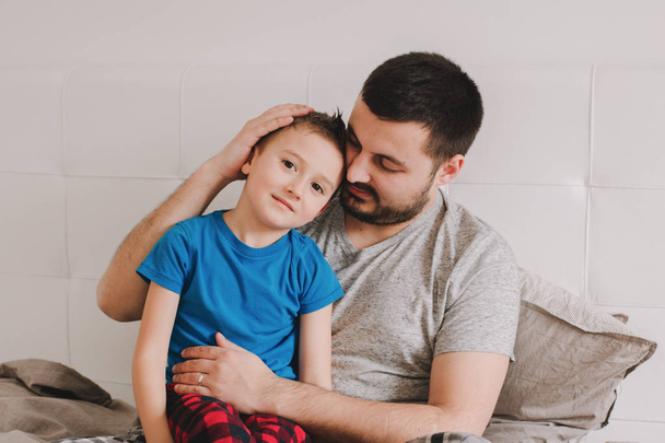Νεαρός Καυκάσιος πατέρας μιλάει στον γιο του. Γονιός που αγκαλιάζεται με παιδί στο κρεβάτι στο σπίτι. Αυθεντικός τρόπος ζωής πραγματική ειλικρινής στιγμή. Ευτυχισμένες διακοπές Ημέρα Πατέρων.  - Φωτογραφία, εικόνα