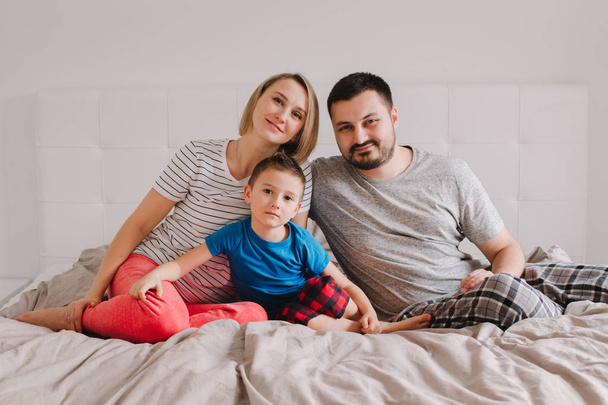 Οικογένεια τριών ατόμων που κάθονται στο κρεβάτι στην κρεβατοκάμαρα κοιτώντας την κάμερα. Μητέρα, πατέρας και γιος στο σπίτι περνούν χρόνο μαζί. Οι γονείς δένονται με το παιδί. Πραγματικοί άνθρωποι αυθεντικός τρόπος ζωής. - Φωτογραφία, εικόνα