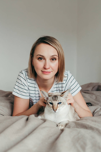 Proud pet owner. Happy Caucasian smiling blonde woman petting oriental cat with blue eyes. Beautiful girl lying on bed in bedroom at home and stroking domestic feline animal.  - Foto, Imagen